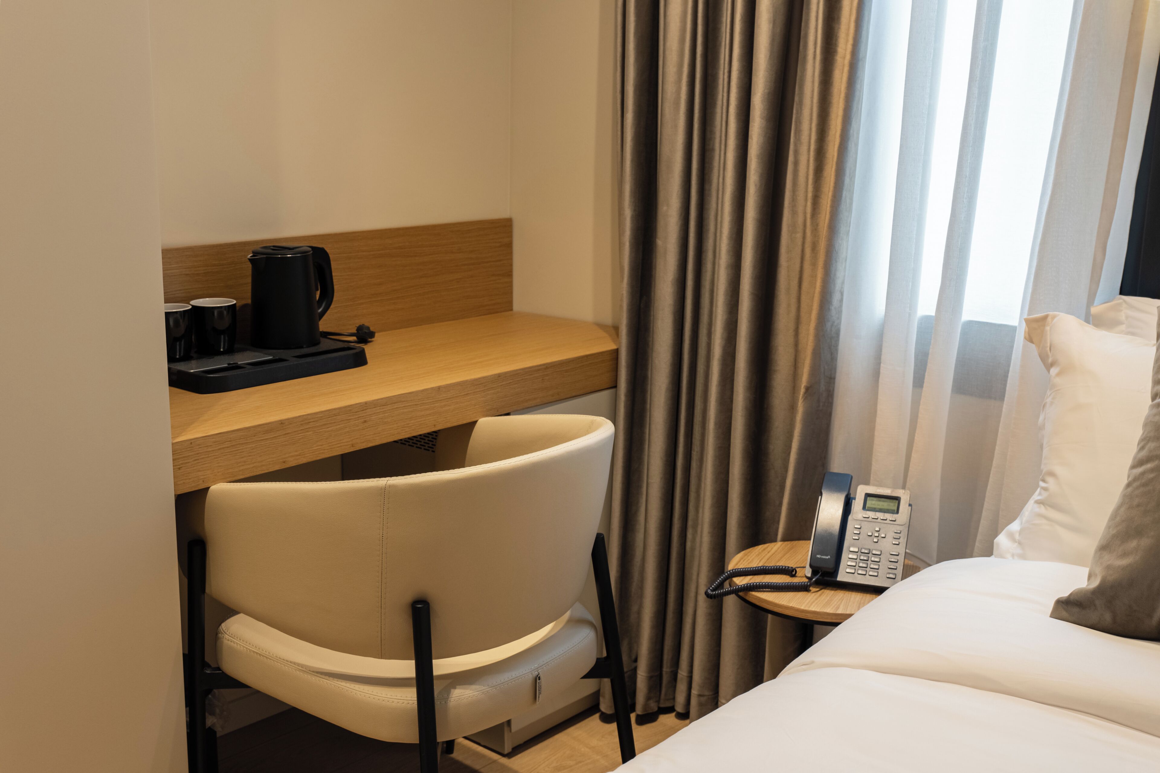 Ein Hotelzimmer mit einem Bett, einem Schreibtisch mit Kaffeemaschine, einem Sessel, einem Telefon und einem Fenster mit Vorhängen.