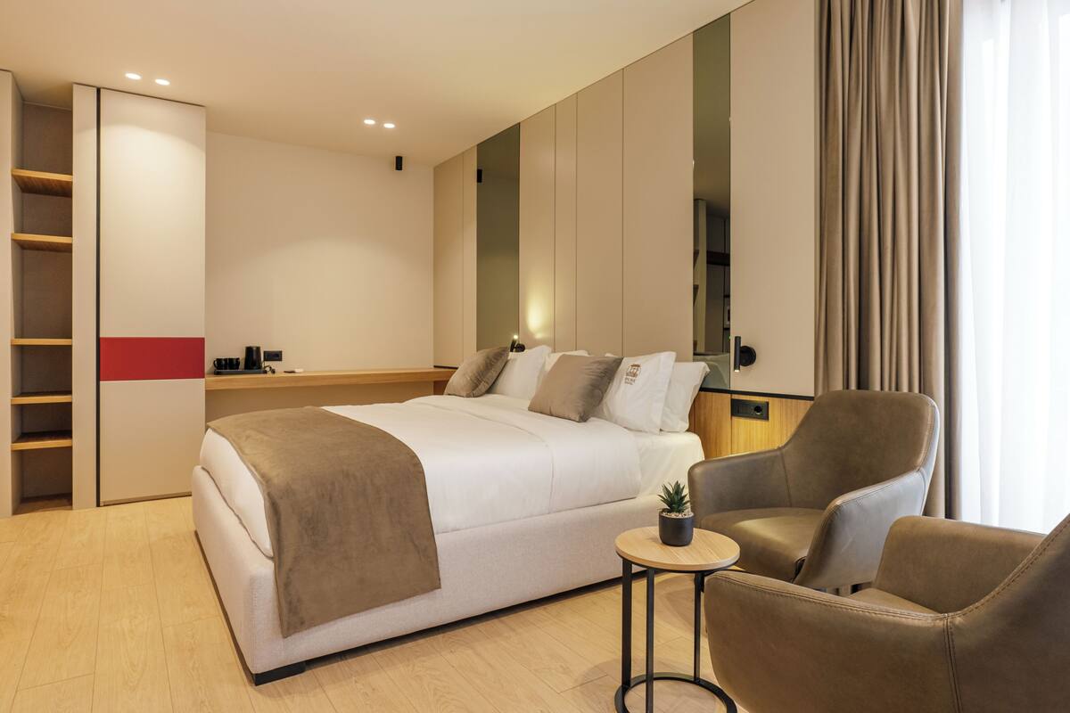A modern hotel room with a bed, a seating area, a small table with a plant, and a wardrobe.