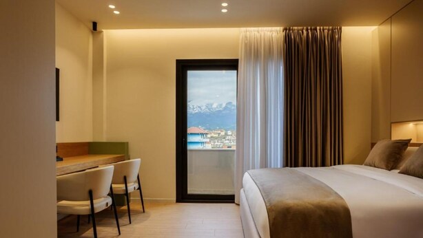A hotel room with a bed, a desk with chairs, a view of mountains through a window, and a balcony. - Pusi Hotel (Tirana)