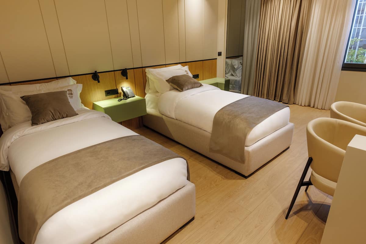 A hotel room with two beds, a nightstand, a chair, and a window with curtains.