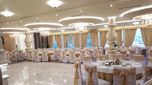 A banquet hall with round tables, white and gold decor, chandeliers, and large windows. - Pusi Hotel (Tirana)