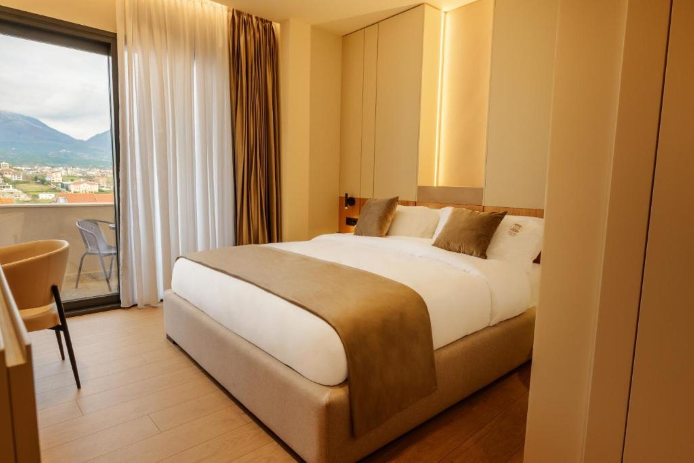 A hotel room with a bed, a chair, a desk, a view of the city, and a balcony.