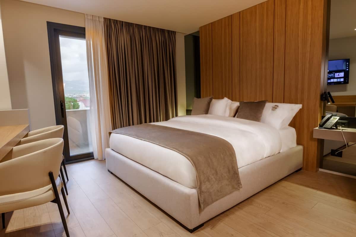 A modern hotel room with a large bed, a desk, a chair, a TV, and a view of the city through a window.
