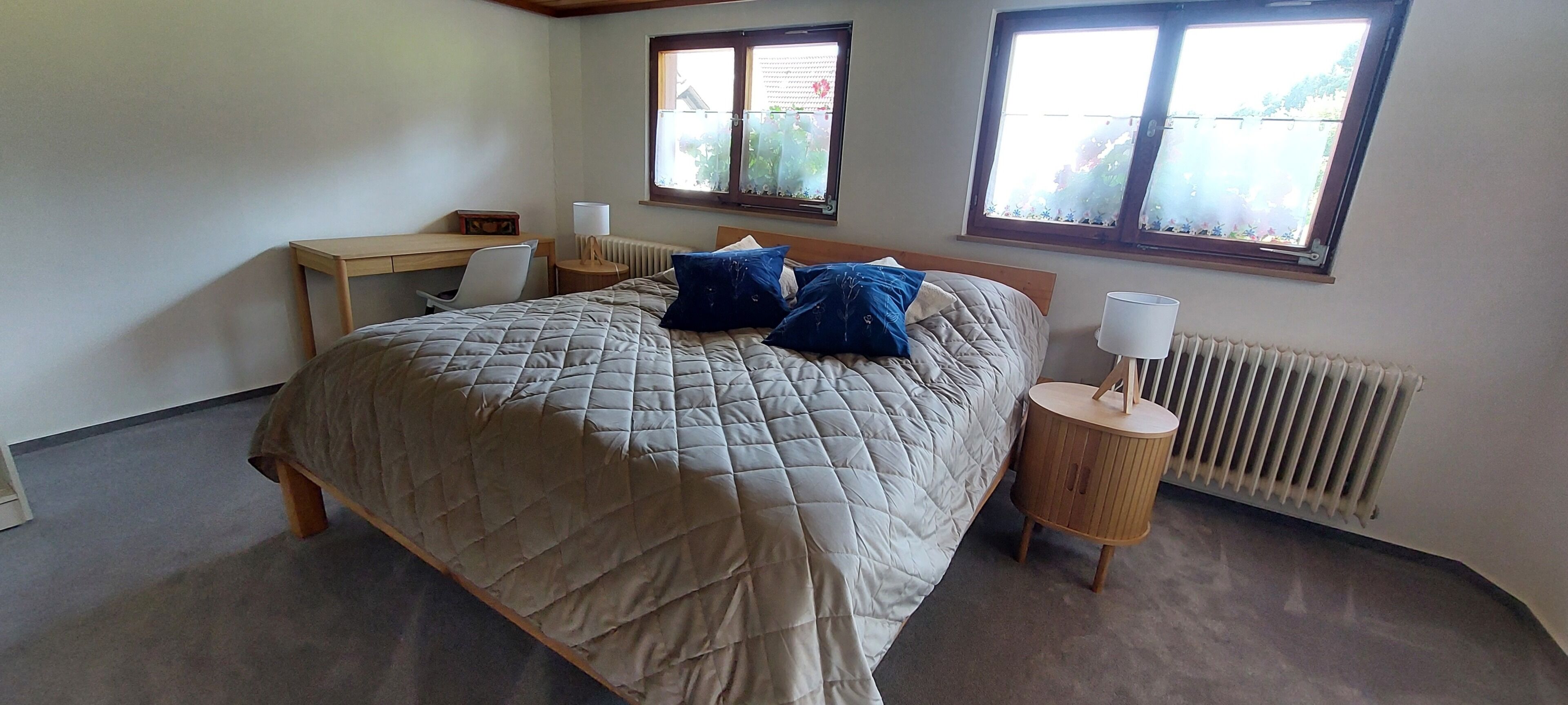 A bedroom with a bed, two windows, a desk, a chair, and a bedside table with a lamp.