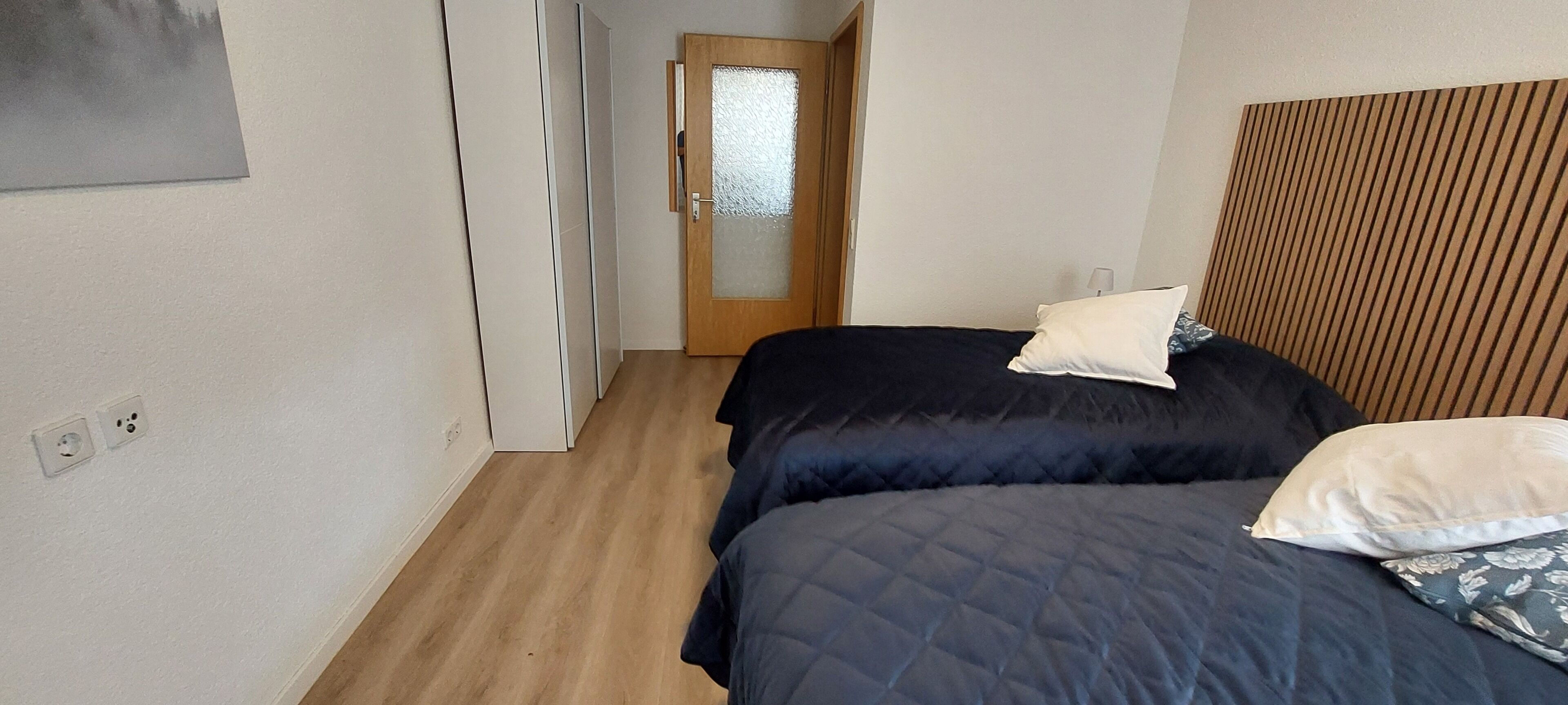 A bedroom with a bed, two pillows, a wooden headboard, a door with frosted glass, and a hallway.