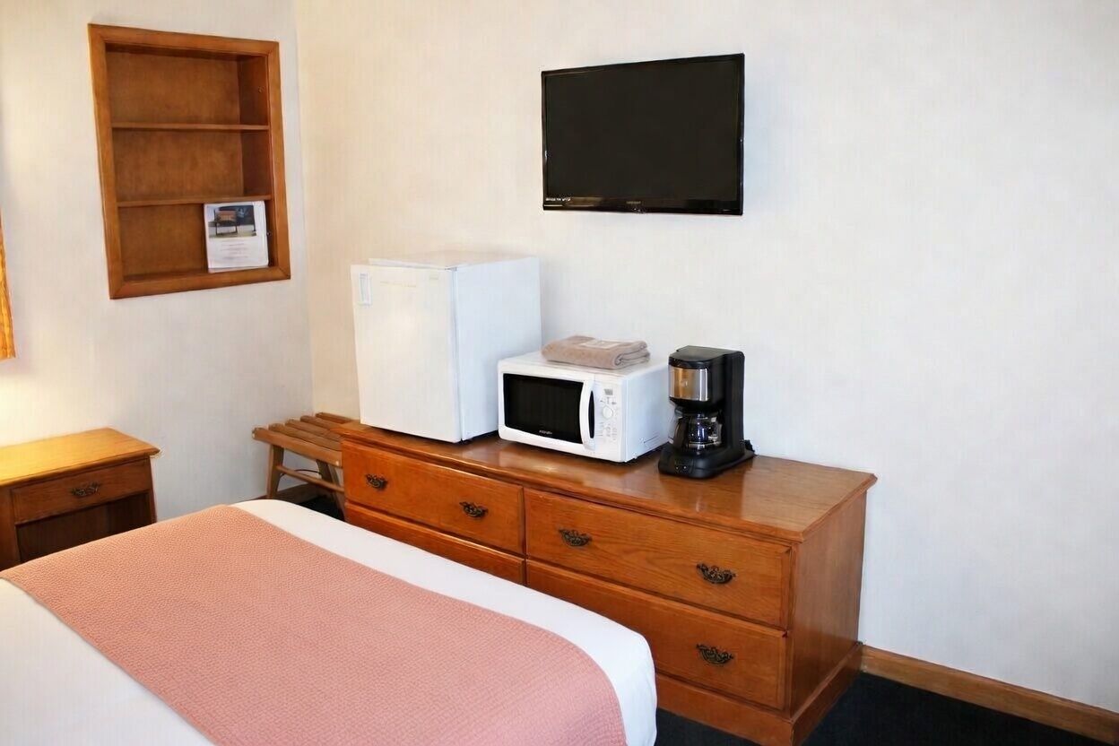 A hotel room with a bed, a wooden dresser, a microwave, a coffee maker, a television, and a small shelf with a picture.