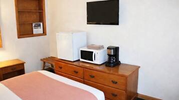 A hotel room with a bed, a wooden dresser, a microwave, a coffee maker, a television, and a small shelf with a picture.