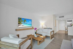 A modern living room with a wooden coffee table, white sofa, and a painting of a beach scene. - Direct Oceanfront + Balcony Suite in 5-Star Resort (Miami Beach)