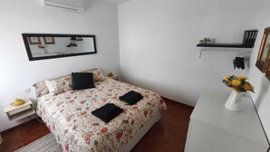 Een slaapkamer met een bed, nachtkastje, dressoir, spiegel, plank met boeken en een vaas met bloemen.