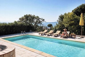 Ein Pool mit Liegestühlen und einem Tisch, umgeben von einer Hecke und Bäumen, mit Blick auf das Meer.