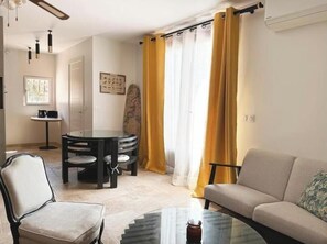 Ein Wohnzimmer mit Sofa, einem Glastisch, einem Esstisch mit Stühlen, einem kleinen Tisch mit Kaffeemaschine und einer Wand-Klimaanlage.
