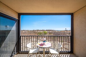 Ein Balkon mit zwei weißen Stühlen und einem Tisch, der auf eine karge Landschaft mit vereinzelter Vegetation und einem strahlend blauen Himmel blickt.