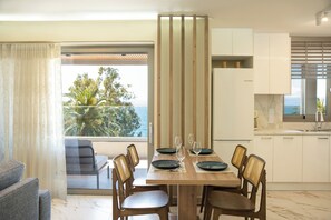 A dining area with a table set for four, a view of the outdoors, and a modern kitchen in the background. - GET Seaside Vibe Apartment Loutraki (Loutraki)