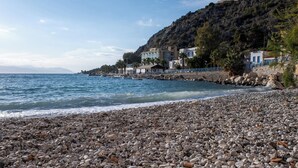 Une plage de galets bordée d’une côte rocheuse, un ciel dégagé et des bâtiments visibles à l’arrière-plan.