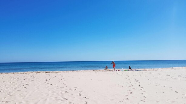 Une vaste plage de sable fin, parsemée d’empreintes menant à l’eau, un ciel bleu azur et une mer calme.