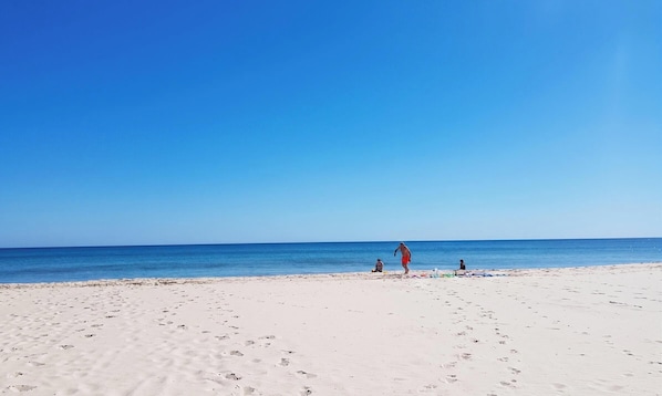 Een breed zandstrand met voetafdrukken die naar het water leiden, een heldere blauwe lucht en een rustige zee.