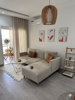 Een moderne woonkamer met een hoekbank, een klein rond tafeltje, een afstandsbediening en een decoratieve hanglamp.