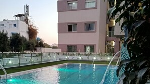 Une piscine agrémentée d’un élément décoratif aquatique, entourée d’une clôture et de verdure, avec un immeuble à plusieurs étages en arrière-plan.