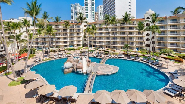 Ein Resortpool mit Liegestühlen und Sonnenschirmen, umgeben von Palmen und Hotelgebäuden.