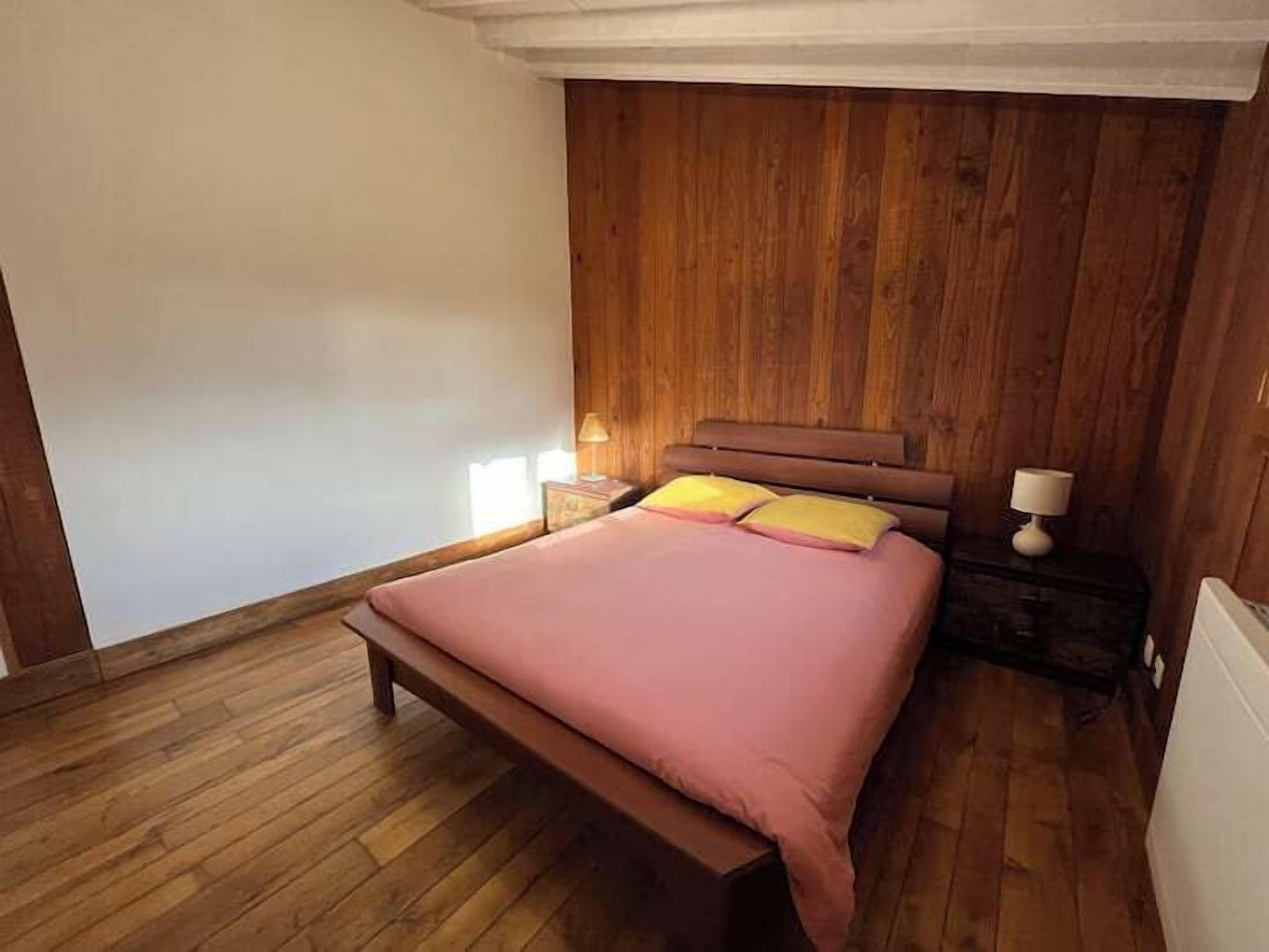 Un dormitorio con cabecera de madera, cama con ropa de cama rosa, mesita de noche con lámpara y suelo de madera.