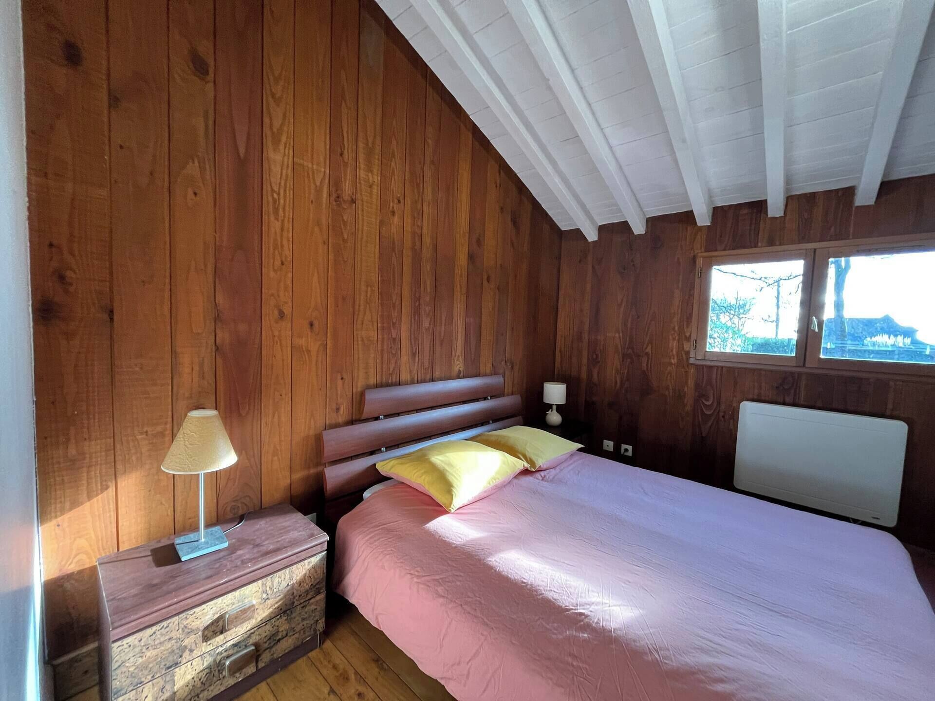 Ein Zimmer mit Holzboden, Bett, Nachttisch, Lampe und Fenster.
