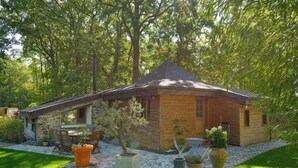 Une petite maison en bois avec une extension en pierre, entourée d’un jardin bien entretenu et d’arbres.