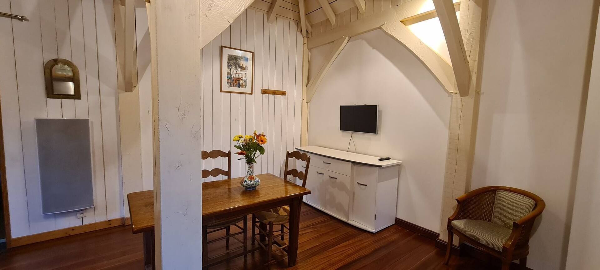 A dining area with a wooden table, chairs, a vase with flowers, a white cabinet, and a television mounted on the wall.