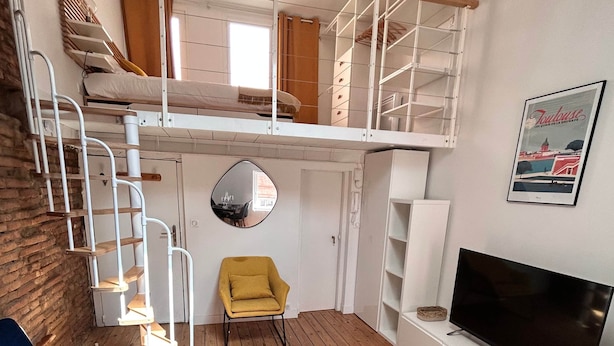 Een kamer in loftstijl met een stapelbed, een televisie, een spiegel en een gele fauteuil.