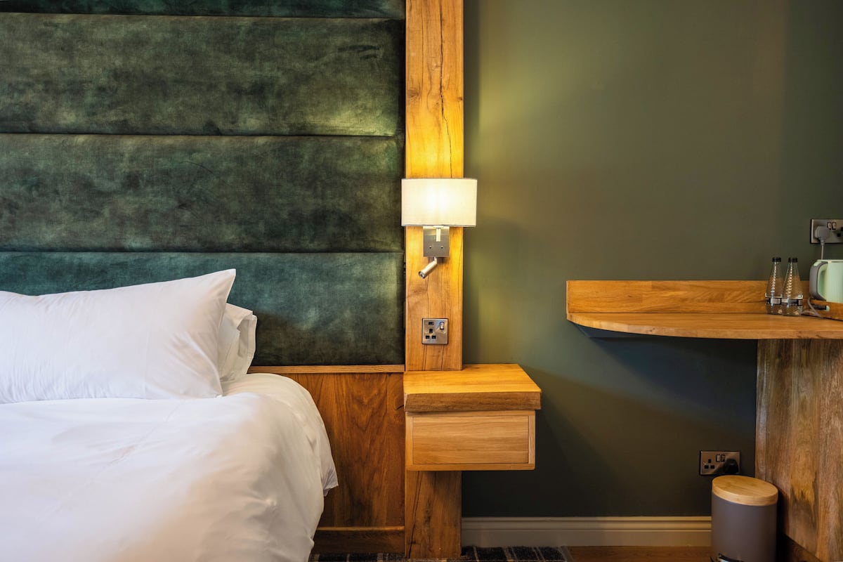 Una habitación de hotel con una cama, una mesita de noche de madera con una lámpara, un tocador de madera con un espejo y un cubo de basura pequeño.
