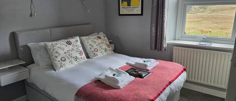 Una cama bien hecha con almohadones florales, una manta roja y un cuadro en la pared.