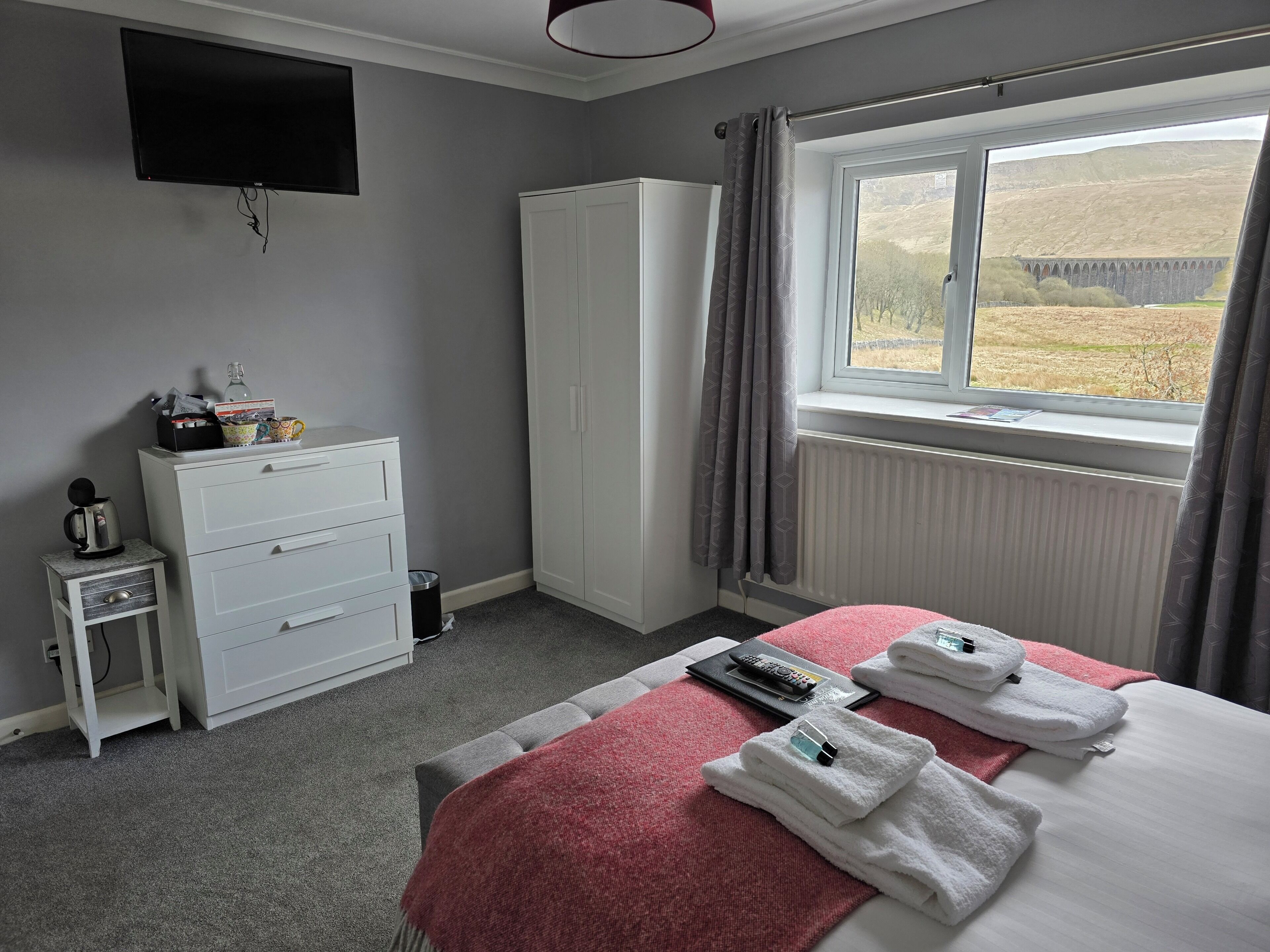 A hotel room with a bed, a TV, a wardrobe, a small table with a kettle, and a window with a view of a bridge.