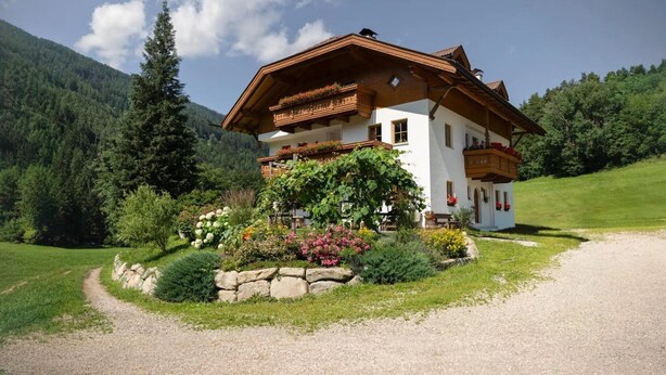 Un chalet de dos pisos con un balcón, rodeado por un jardín con diversas plantas y flores, ubicado en una zona montañosa.