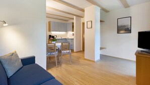 Una sala de estar moderna con un sofá, una mesa de comedor con sillas, una cocina al fondo y un televisor de pantalla plana sobre un mueble de madera.