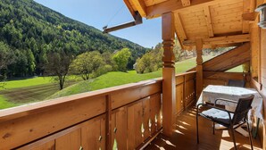 A wooden balcony with a table and chairs, overlooking a green landscape and forested hills. - Apartment 'Getzenbergerhof' with Balcony and Wi-Fi (Chienes)