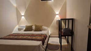A small, cozy bedroom with a bed, patterned pillows, a bedside table, a chair, and a small rug. - Hostal Caimán del Rio (Barranquilla)
