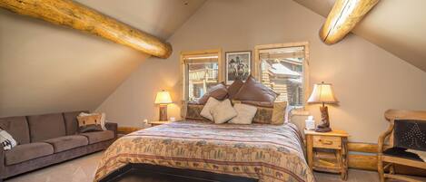 A bedroom with a sloped ceiling, wooden beams, a large bed with patterned bedding, a nightstand with a lamp, a wooden chair, and a sofa.