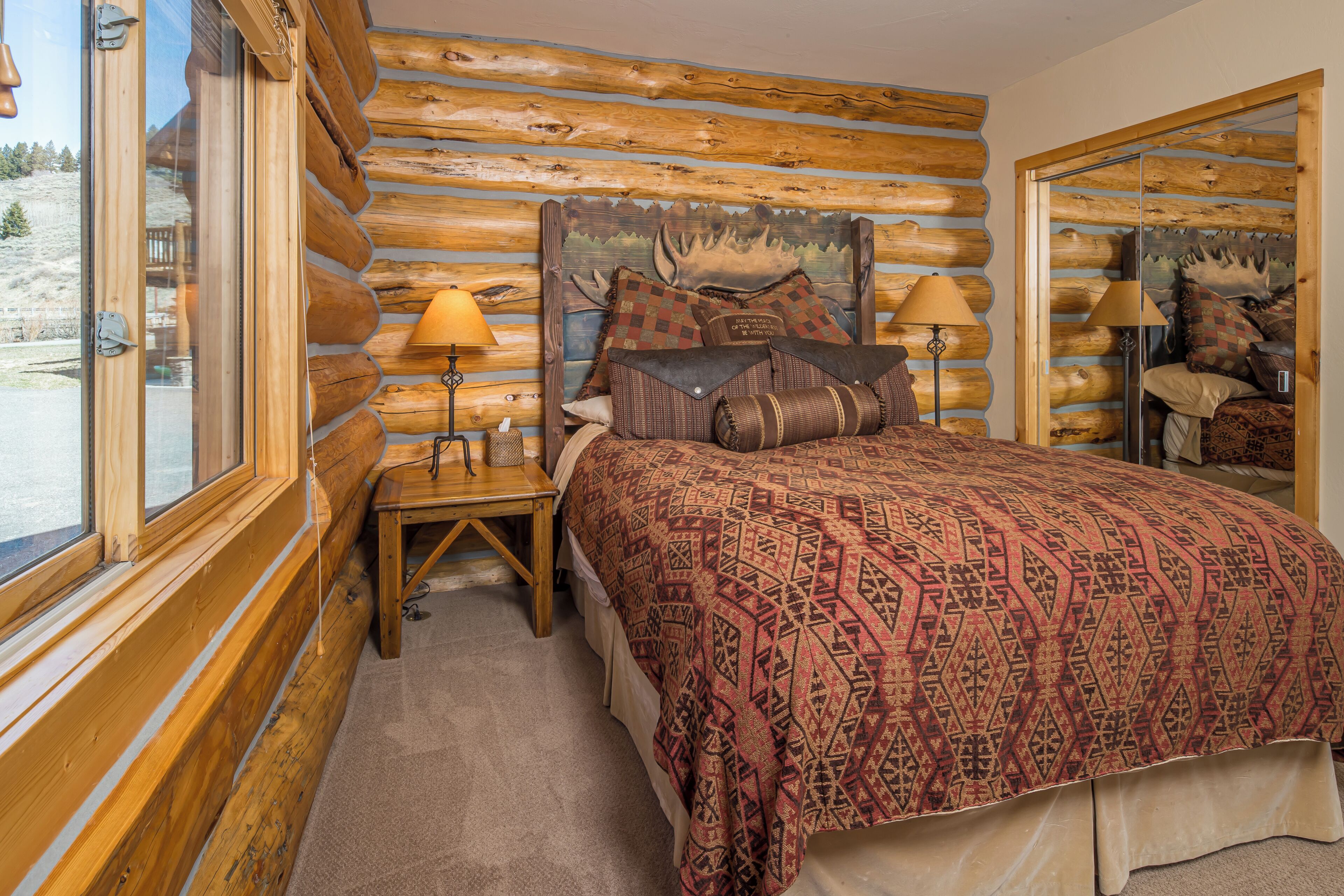 A log cabin-style bedroom with a bed, two bedside tables, lamps, and a large window.