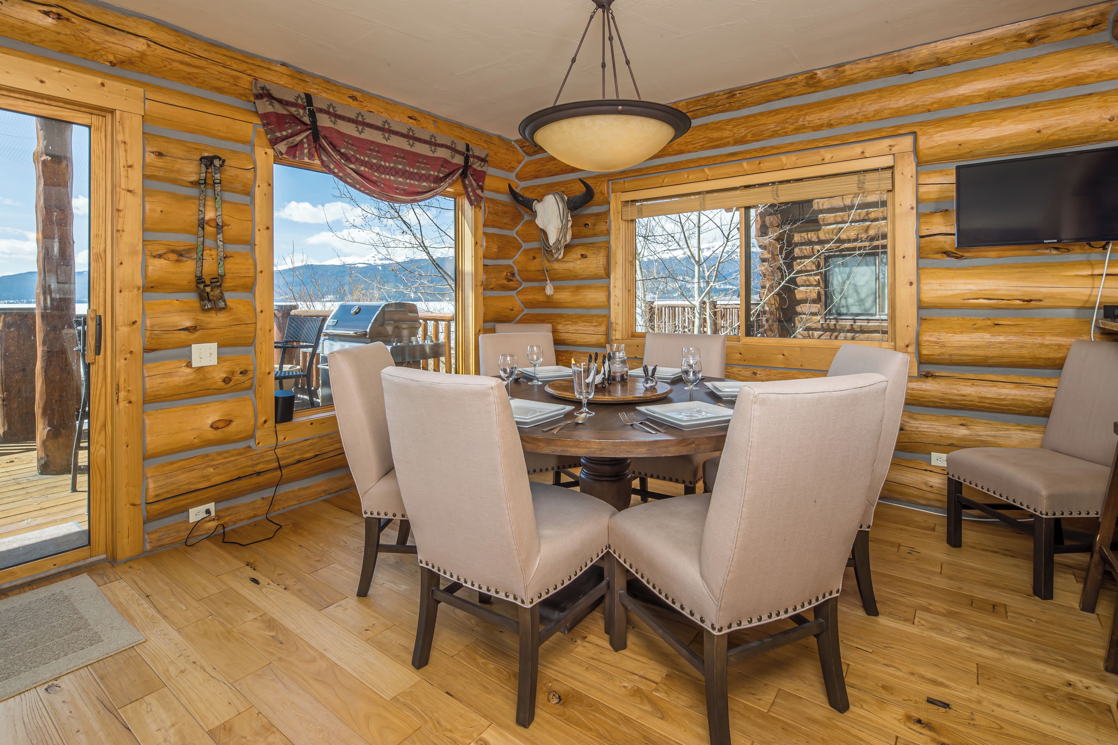 A wooden cabin interior with a dining table set for four, chairs, a mounted television, and a large window with a mountain view.