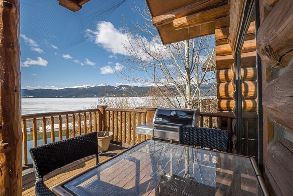 Eine Holzterrasse mit Grill, Stühlen und einem Glastisch mit Blick auf einen See und Berge.
