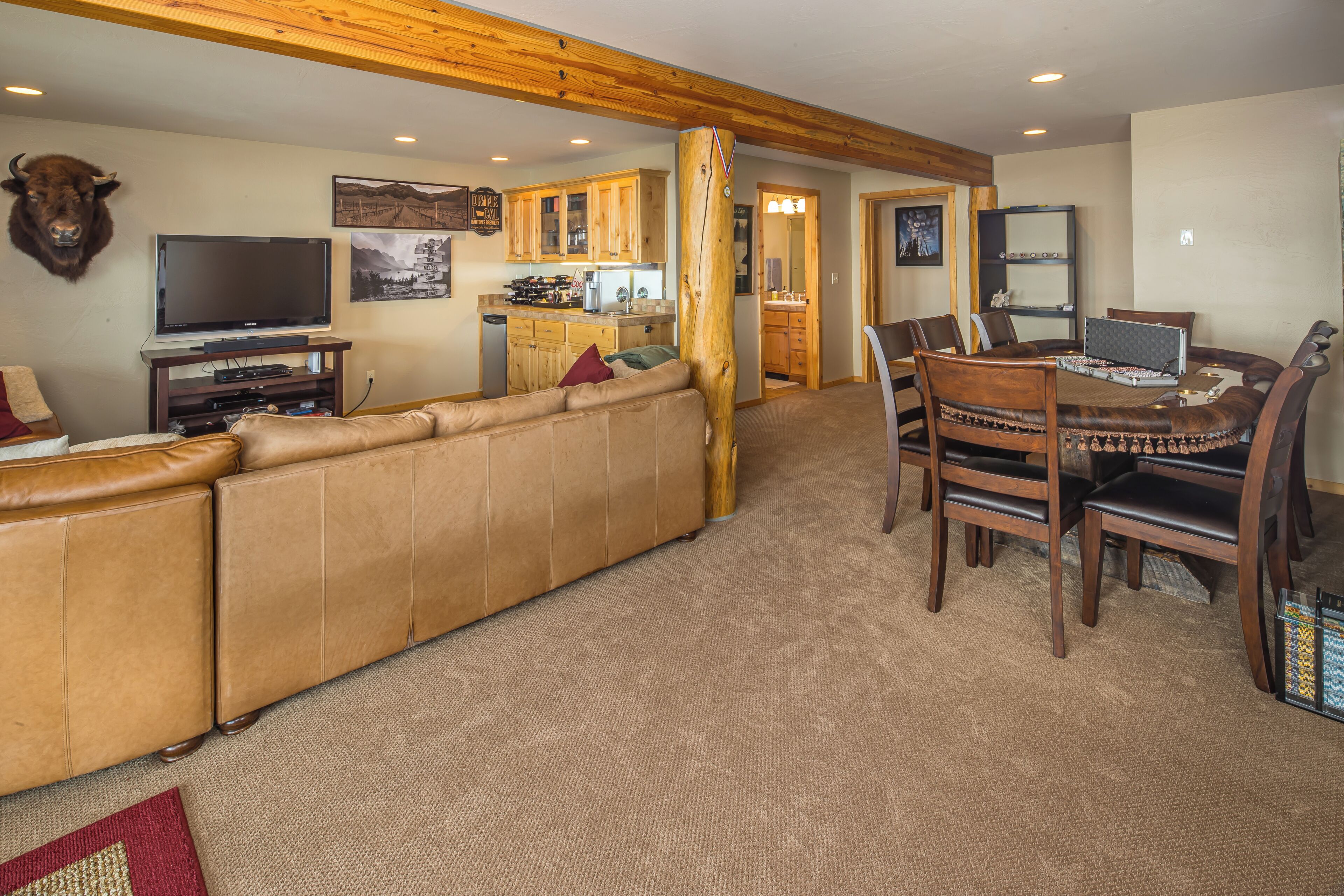 A living room with a large leather sofa, wooden dining table, and chairs, a kitchen in the background, and a mounted animal head on the wall.