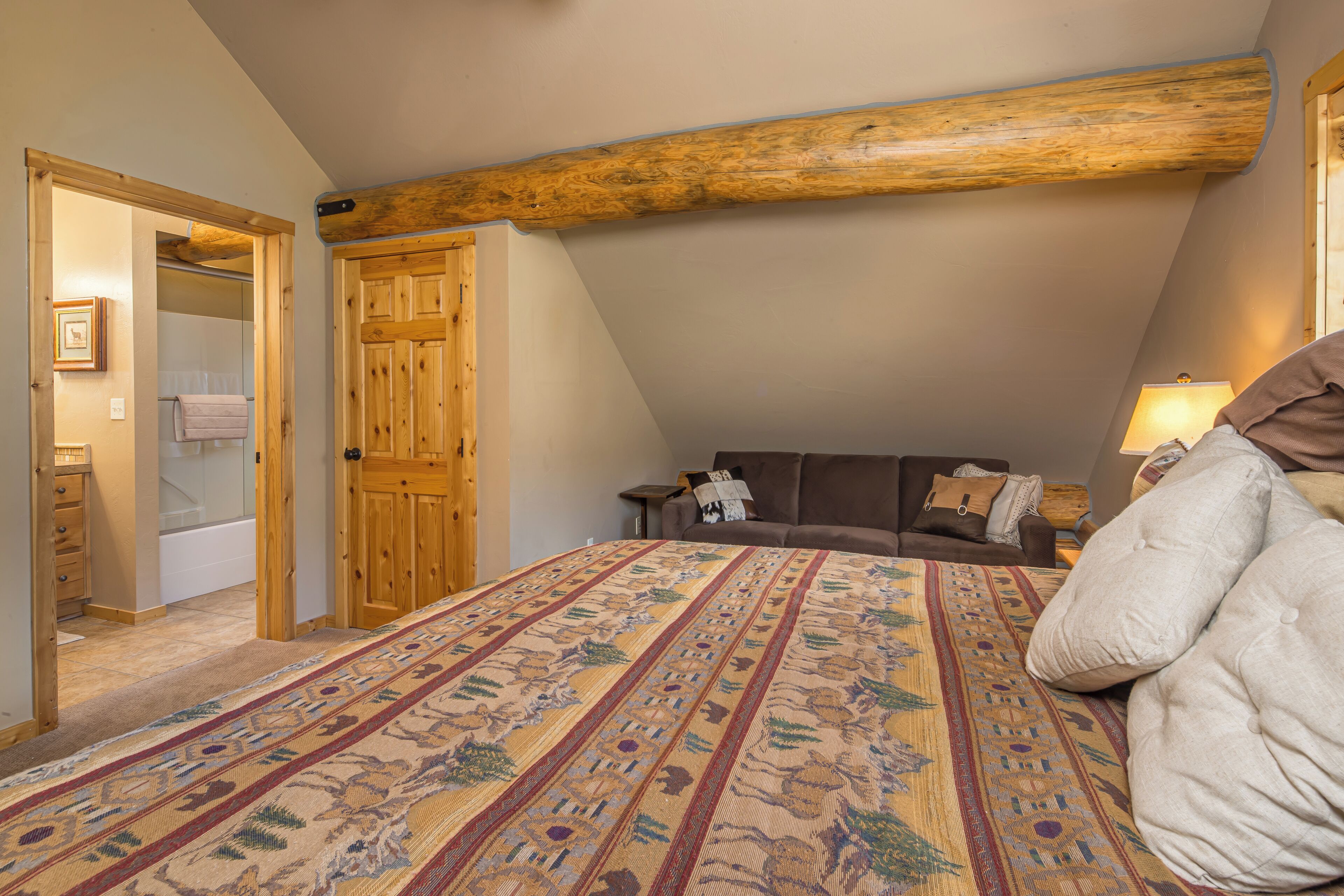 A bedroom with a large bed, a wooden ceiling beam, a door, a nightstand, and a lamp.