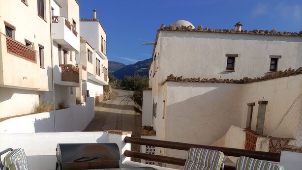 An outdoor patio with a table and striped chairs, overlooking a street with buildings and a mountain in the distance.