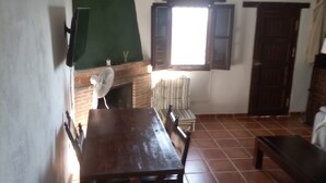 A dining area with a wooden table and chairs, a green wall, a window with open shutters, and a tiled floor.