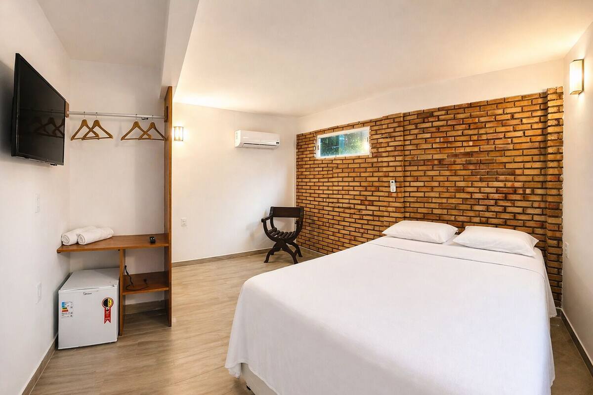 A hotel room with a brick accent wall, a bed, a desk with a chair, a TV, and a small refrigerator.