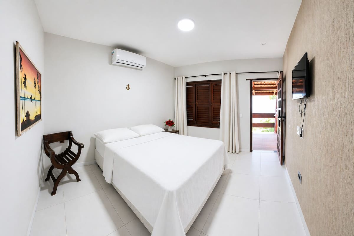 A bedroom with a bed, a chair, an air conditioning unit, a TV, and a painting on the wall.