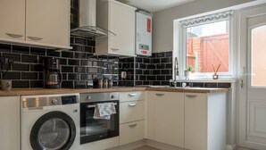 A compact kitchen with a washing machine, oven, and a window with a view of a red fence. - Stylish house with Backyard + 4 min walk to Beach (Great Yarmouth)