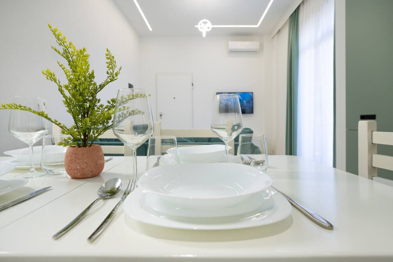 A dining area with white table settings, including plates, cutlery, and wine glasses. A potted plant is on the table, and there is a painting on the wall.