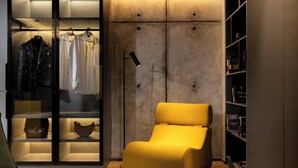 Una stanza con una poltrona gialla, un guardaroba con sportello in vetro e abiti appesi, e una libreria.