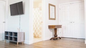 A room with a flat-screen TV mounted on the wall, a wooden sideboard with a shelf, a small wooden table, and a white door.