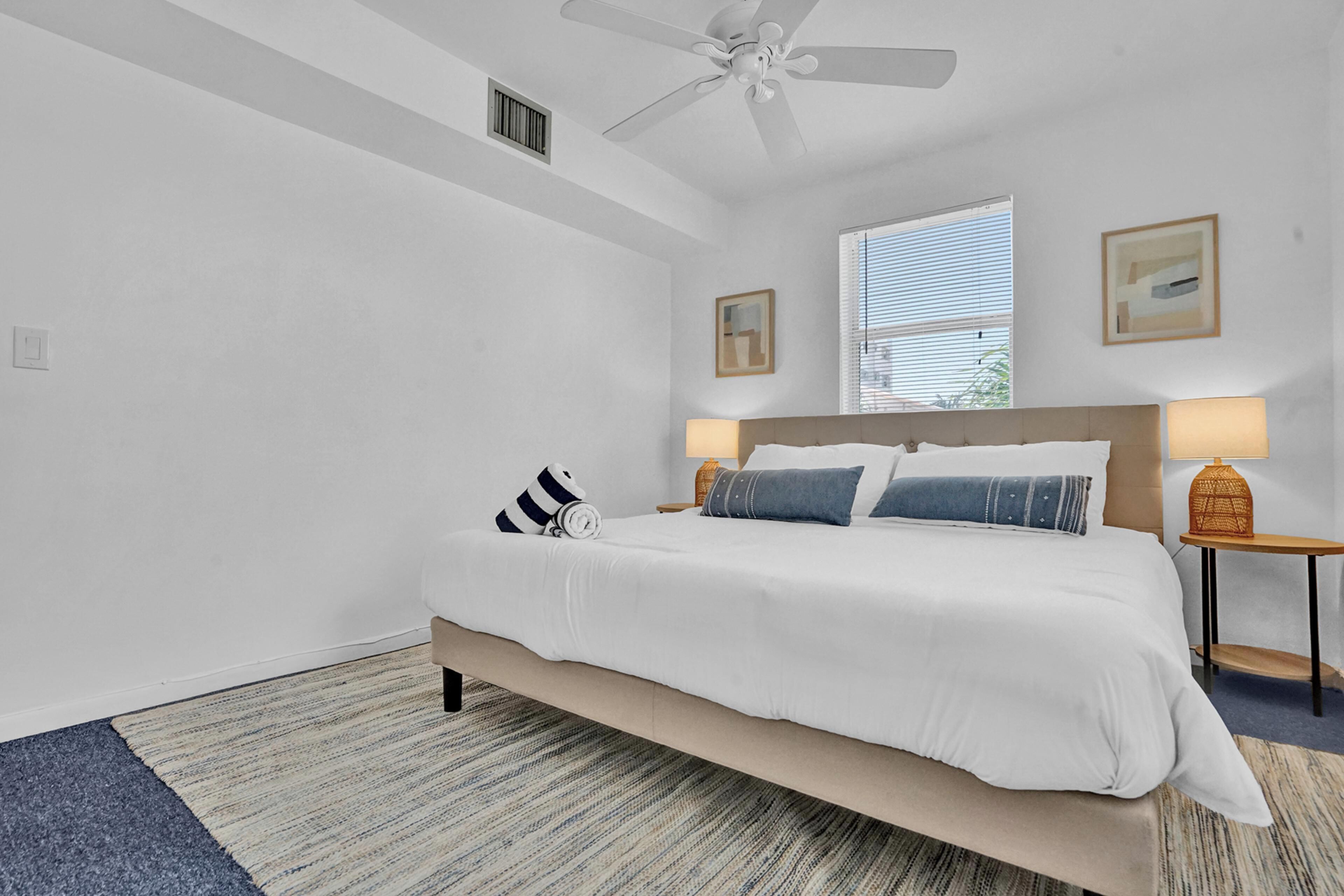 A bedroom with a bed, a ceiling fan, a window with blinds, and two wall-mounted lamps.